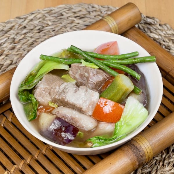 A white bowl of Filipino Sinigang soup with pork, long beans, bok choy, tomatoes, and eggplant.