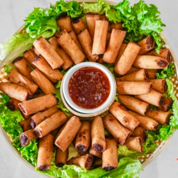 A platter of crispy spring rolls arranged in a circle around a bowl of dipping sauce, on a bed of lettuce.